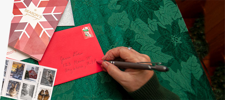 Person writing an address on a holiday card envelope featuring a holiday stamp.