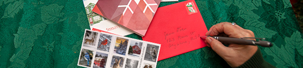 Person writing an address on a holiday card envelope featuring a holiday stamp.